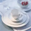 A white porcelain breakfast plate with a cup and saucer on top, set on a white placemat. Produktbild-Vorschau