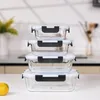 A stack of four glass food containers with black and white lids on a white surface, with a blurred kitchen background. Náhled obrázku produktu