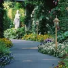 A winding garden path lined with flowers and greenery, featuring two ornate lampposts and a classical statue. Podgląd zdjęcia produktu