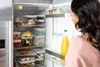 A woman is looking into an open refrigerator filled with food containers and bottles. Náhľad obrázku produktu