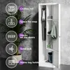 A white metal locker with its door open, showcasing its interior features. The locker is placed against a textured gray wall, with a large potted plant to its left. Various icons and text labels highlight the locker's features. Náhled obrázku produktu