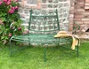 A green metal garden bench sits on grass in front of a stone wall with a rose bush. Náhľad obrázku produktu