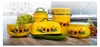 A collection of yellow ceramic kitchenware with sunflower designs, including canisters and a butter dish, arranged on a wooden countertop. Green and red grapes are placed around the items. Produktbild-Vorschau