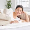 A woman is lying in bed, smiling and holding a heating pad controller. Náhľad obrázku produktu