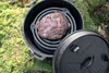 A cast iron Dutch oven with a piece of meat inside, sitting on mossy ground. Produktbild-Vorschau