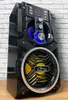 A black portable Bluetooth speaker with LED lights and a yellow speaker cone is shown against a white brick wall. Produktbild-Vorschau