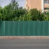 A green corrugated metal fence runs along a paved road, with trees and a building in the background. Voir les images du produit
