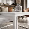 A close-up of a white high-gloss coffee table in a living room setting, with pillows and a vase in the background. Produktbild-Vorschau