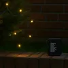 A string of warm white LED lights is draped over a Christmas tree branch, with the battery pack resting on a brick ledge. Produktbild-Vorschau
