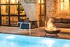A fire pit is placed next to a swimming pool, with outdoor furniture and a stone building in the background. Voir les images du produit