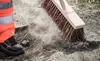 A person in high-visibility work pants and safety shoes is sweeping dirt and dust with a broom on a rough, outdoor surface. Produktbild-Vorschau
