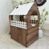 A wooden cat house with white accents, placed on a textured surface with decorative white flowers in the background. Produktbild-Vorschau