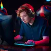A young man wearing a gaming headset is playing a video game on a computer in a dimly lit room with colorful lighting. Produktbild-Vorschau
