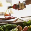 A hand is pouring Hennessy VSOP cognac from a bottle into a glass, with books and vegetables in the foreground. Produktbild-Vorschau