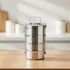 A stainless steel food container with four stacked compartments sits on a wooden table in a kitchen setting. Produktbild-Vorschau