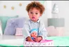 A young child is holding a baby doll in a carrier, surrounded by soft furnishings. Produktbild-Vorschau
