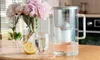 A water filter pitcher and a glass of water are on a wooden table, with a bouquet of pink peonies in the background. Náhled obrázku produktu