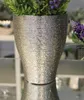 A close-up shot of a silver textured vase with green and purple artificial plants inside, placed on a reflective surface. Produktbild-Vorschau