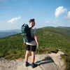 A man stands on a rocky outcrop overlooking a vast, green, forested mountain range under a clear blue sky with scattered clouds. Anteprima dell’immagine prodotto