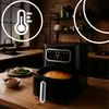An air fryer is shown on a wooden countertop in a kitchen setting, with food on a plate and a bowl of sauce nearby. There are also graphic overlays indicating temperature and another circular graphic. Produktbild-Vorschau