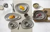 A collection of taupe-colored ceramic tableware, including plates, bowls, and cups, arranged on a light-colored textured surface. Produktbild-Vorschau