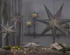 A collection of grey paper star lanterns are displayed in a dimly lit room, some hanging and others on a shelf and table, with fairy lights and greenery. Podgląd zdjęcia produktu