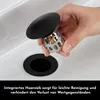 A hand is shown holding a black drain stopper with an integrated hair sieve over a sink drain. Produktbild-Vorschau