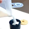 A close-up shot of white granular material being poured from a clear plastic container into a dark blue ceramic container with a wooden lid. Anteprima dell’immagine prodotto