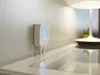 A white Philips Hue Bridge is mounted on a wall above a white surface, with a lamp and a small plant in the background. Produktbild-Vorschau