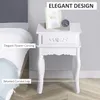 A white nightstand with a drawer, decorated with floral carvings, placed next to a bed. Produktbild-Vorschau
