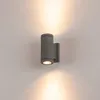 A gray cylindrical outdoor wall light is mounted on a light gray wall, emitting light upwards and downwards. Produktbild-Vorschau