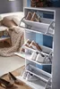 A white shoe cabinet with multiple shelves filled with shoes, placed in a bedroom setting next to a bed with blankets and pillows. Produktbild-Vorschau