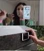 A woman is applying makeup in front of a mirror, with a smartphone displaying a makeup app. The second image shows a hand holding a smartphone against a stone surface. Produktbild-Vorschau