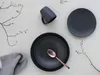 A flat lay of dark gray stoneware plates, a mug, and a rose gold spoon, arranged with decorative branches and a gray napkin on a light gray background. Produktbild-Vorschau