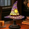 A witch hat-shaped stained glass lamp sits on a wooden table next to a cup of coffee and a stack of books. Produktbild-Vorschau
