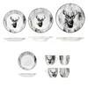 A collection of ceramic tableware, including plates, bowls, and cups, featuring a black and white stag motif against a wood-grain background. Voir les images du produit