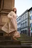 A woman walks down stone steps in a city street, wearing a long beige coat and a matching hat. Produktbild-Vorschau
