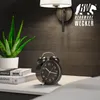 A bedside table with an alarm clock, books, and a plant, illuminated by a wall lamp. Produktbild-Vorschau