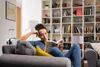 A woman is reading a book on a couch in a living room with a bookshelf in the background. Produktbild-Vorschau