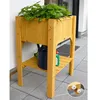 A wooden raised garden bed with plants, a watering can, and a bottle of plant food on a tiled patio. Produktbild-Vorschau