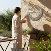 A woman is standing next to a stone wall, interacting with a sundial. There are plants and a table with a book and glasses in the foreground. Produktbild-Vorschau