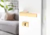 A close-up of a gold-colored door handle and lock set on a white door, with a blurred background of a living room. Produktbild-Vorschau