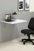 A white folding desk mounted on a gray wall, with a laptop and a lamp on it. A black office chair is in the foreground. Produktbild-Vorschau