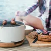 A person is grilling meatballs on a portable grill by the water, with bread and a knife on a cutting board nearby. Produktbild-Vorschau