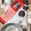 A flat lay of baking ingredients including a bar of Manner chocolate, chopped chocolate, eggs, milk, flour, and a whisk. Produktbild-Vorschau