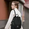 Schwarzer Stoffrucksack mit silbernem ineinandergreifendem Logo, getragen von einer Frau in weißem Mantel, die im Freien geht Produktbild-Vorschau