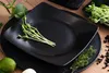 A close-up overhead view of two black square plates, one stacked on top of the other, with microgreens placed on the top plate. There is also a lime and some herbs in the foreground. Náhled obrázku produktu