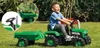 A young boy is happily riding a green pedal tractor with a trailer in a grassy yard. Produktbild-Vorschau