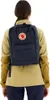 A person wearing a tan and brown jacket is shown from the back, wearing a navy blue backpack. Produktbild-Vorschau
