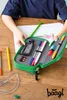 A child's hands are shown opening a green pencil case filled with pens, pencils, and erasers, on a wooden desk with a drawing and a ruler. Produktbild-Vorschau